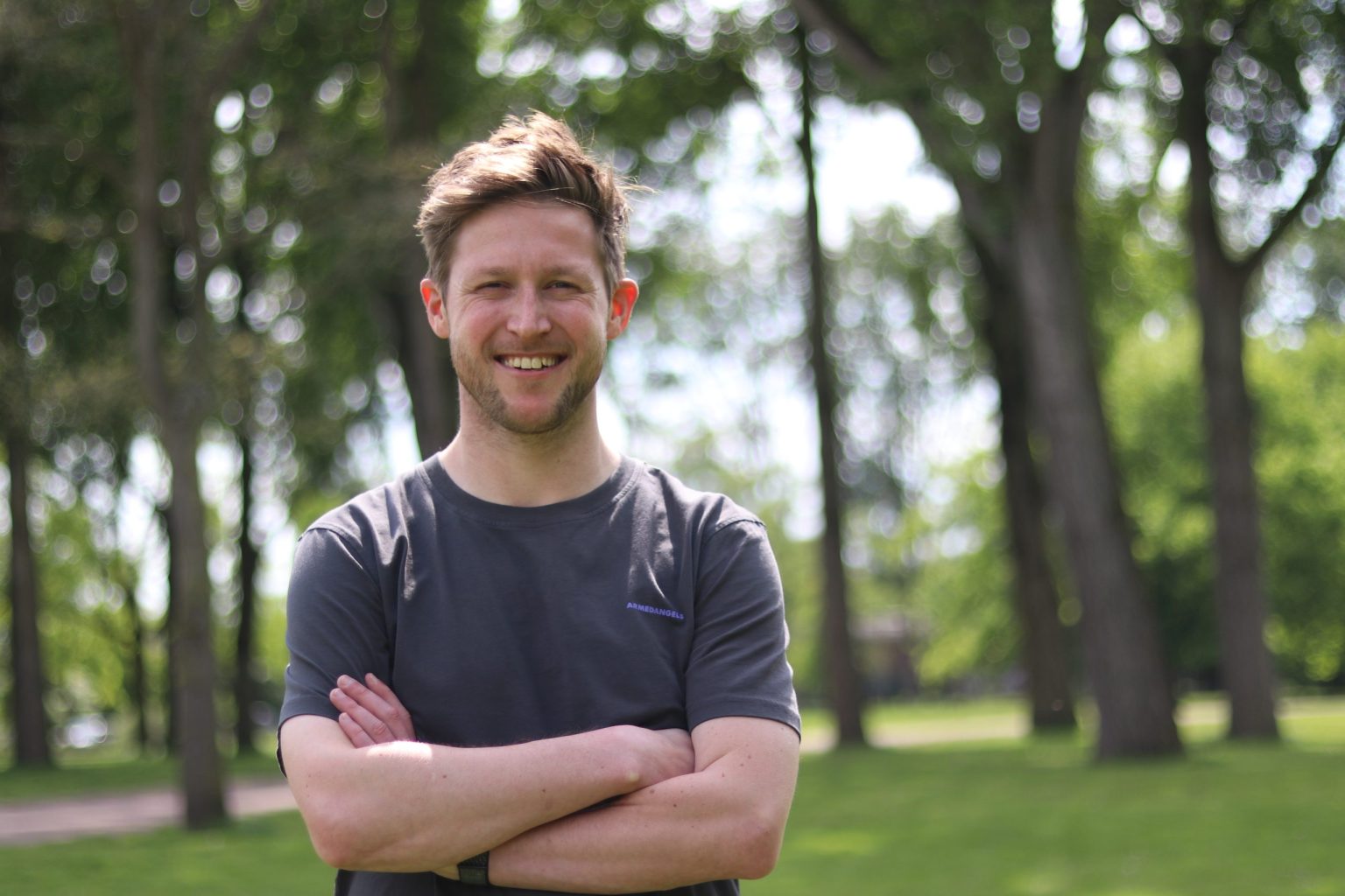Bram staat met armen over elkaar buiten in een park, glimlachend naar de camera.